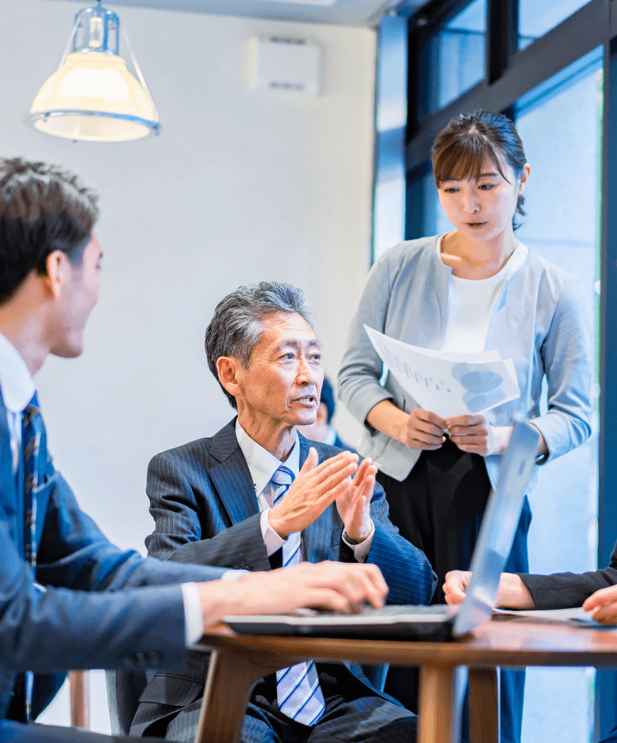 会議中の風景。年配の男性がプレゼンをしながら、周囲の若いスタッフと話し合っている。女性スタッフが資料を持ちながら補足説明をしている。