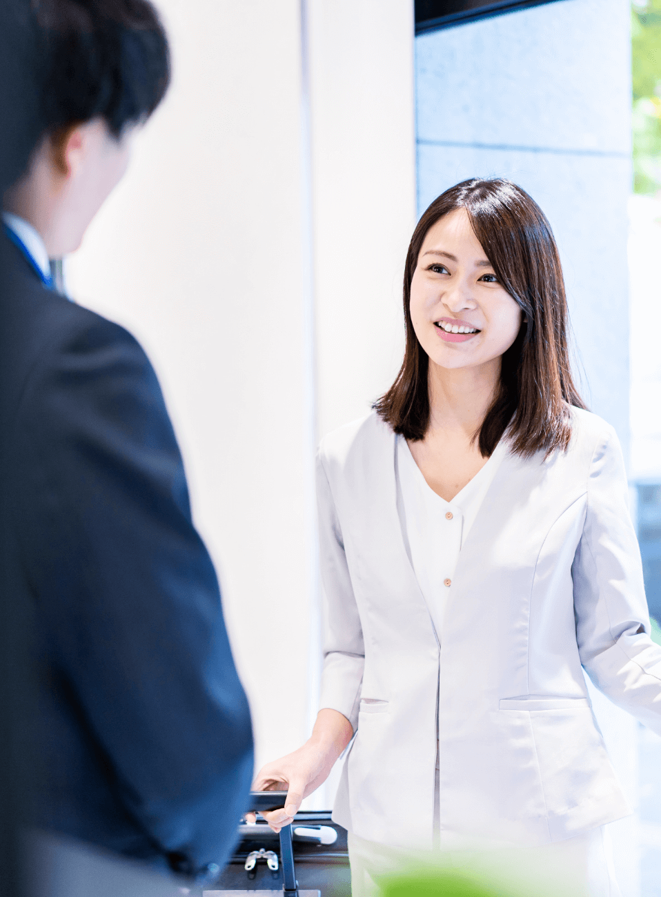 白い制服を着た女性が、来訪者と微笑みながら会話している。明るい受付でのシーンで、親しみやすく丁寧な接客の様子が伝わる。